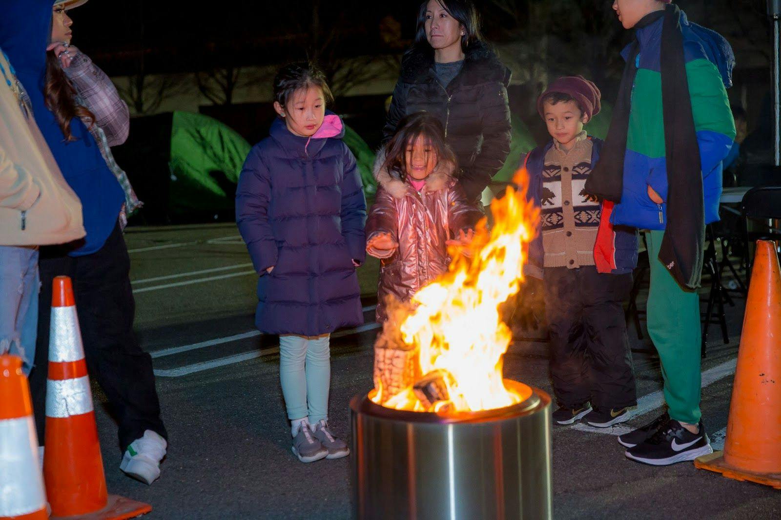 A group of children and adults gathers around a fire in a barrel in an Eastern Mountain Sports parking lot. 